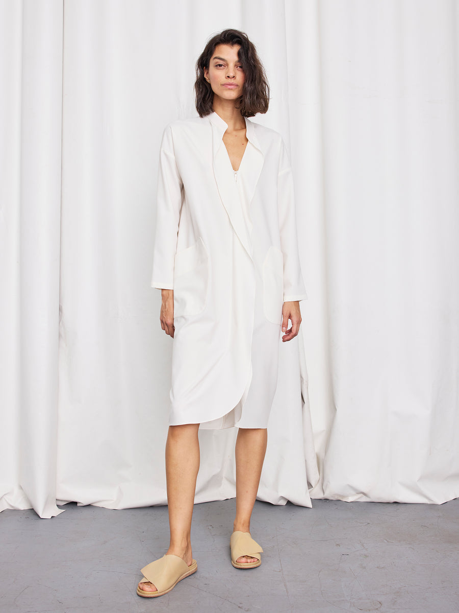 A person with short dark hair stands before white curtains, wearing the Zero + Maria Cornejo Long-Sleeved Fin Gaban Dress and beige slides, embodying effortless style in this New York-made look.