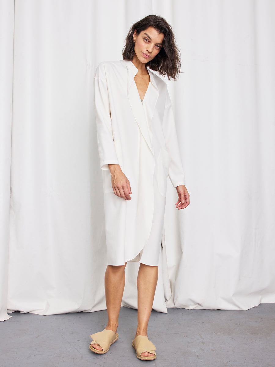 A person with shoulder-length hair wears the Zero + Maria Cornejo Long-Sleeved Fin Gaban Dress and beige sandals, standing in front of white curtains on a gray floor.