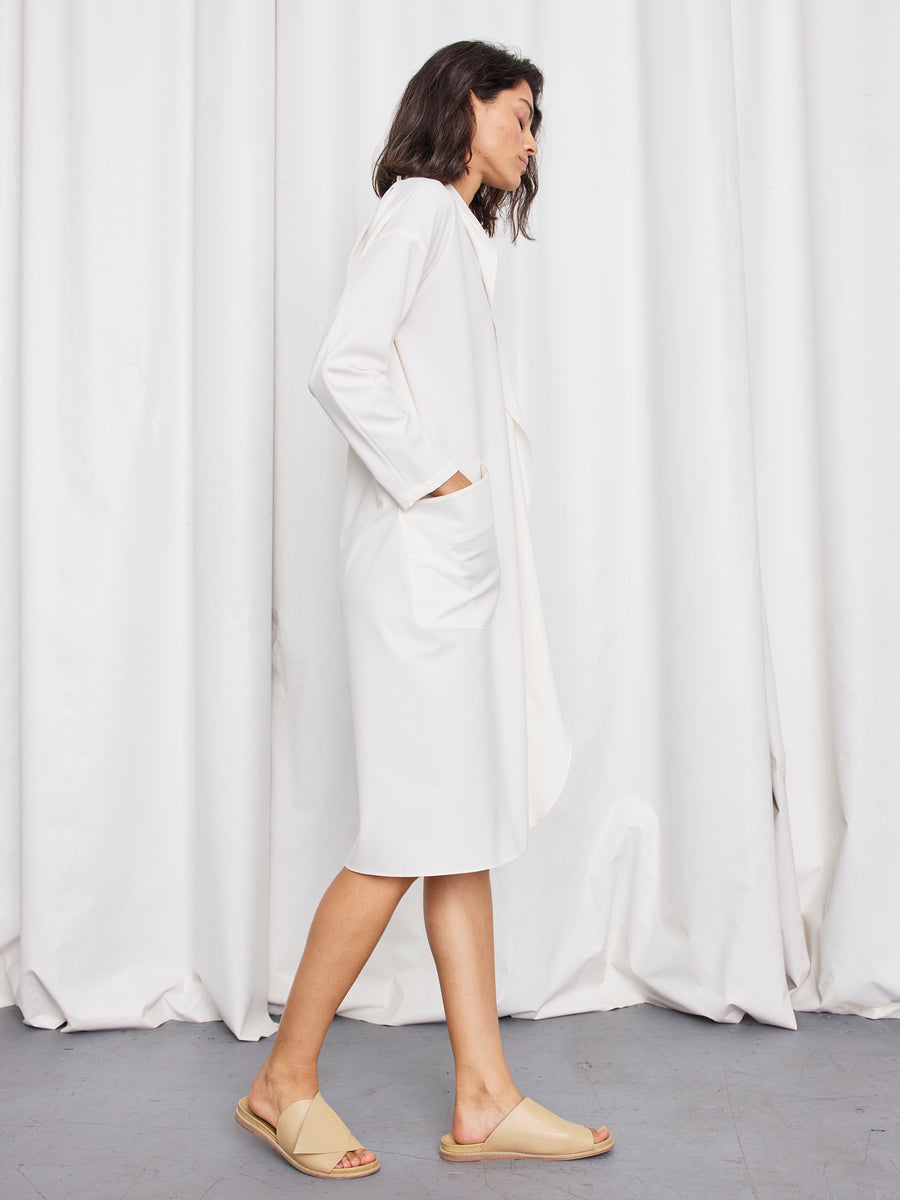 A woman stands in profile against white curtains, hands in her pockets, wearing the Zero + Maria Cornejo Long-Sleeved Fin Gaban Dress and beige slide sandals—a chic style crafted in New York.