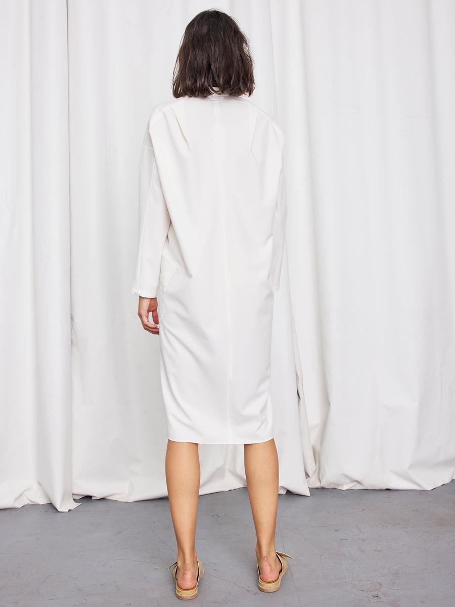 A woman stands barefoot on a gray floor, facing away in the Long-Sleeved Fin Gaban Dress by Zero + Maria Cornejo. Loose-fitting with a draped collar, this elegant knee-length dress is made in New York. White curtains hang in the background.