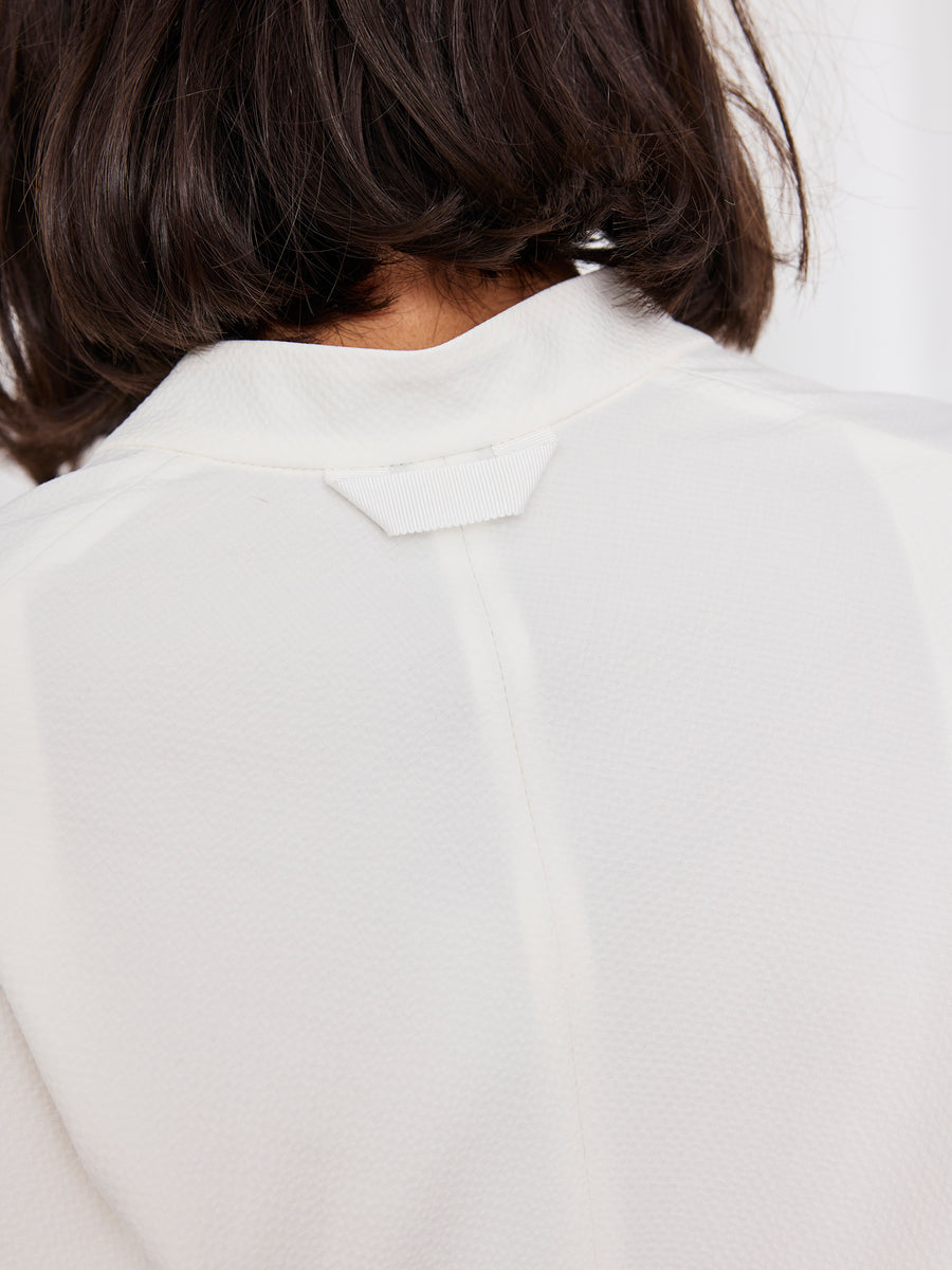 A close-up shows the back of a person in the Zero + Maria Cornejo Long-Sleeved Fin Gaban Dress. Their dark bob partly covers the white textured collar, highlighting the dress’s elegant and relaxed style at the upper shoulders and neck.