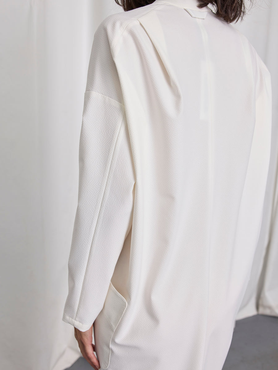 A person with dark hair, seen from behind, wears the Zero + Maria Cornejo Long-Sleeved Fin Gaban Dress—off-white with textured fabric, a back seam, and a subtle draped collar—standing before white curtains.