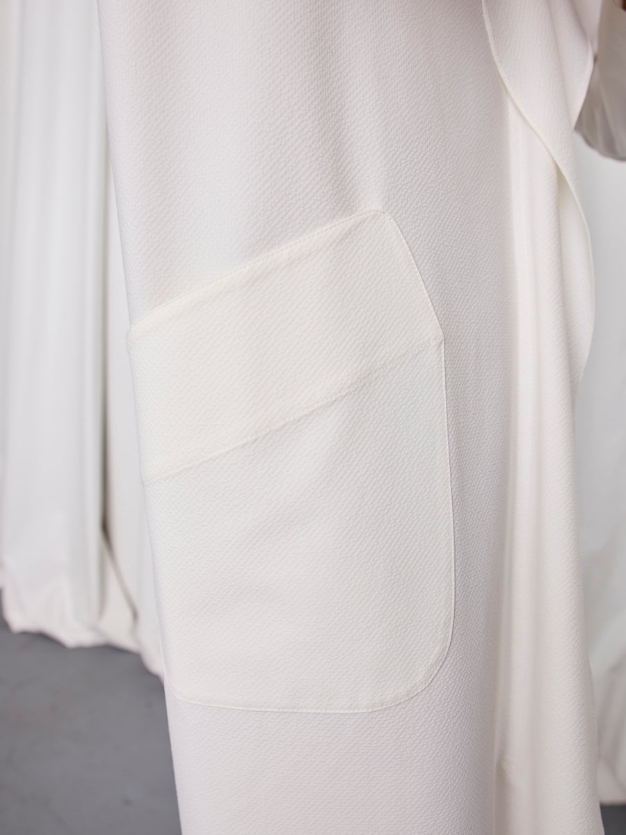 Close-up of the textured cream fabric on the Long-Sleeved Fin Gaban Dress by Zero + Maria Cornejo, featuring a large square pocket. Matching draped fabric and a gray floor in the background emphasize its elegant design.