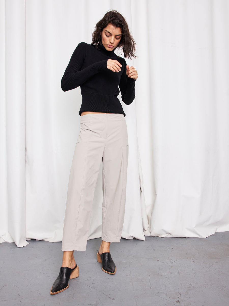 A woman stands indoors by white curtains, wearing a black turtleneck, Zero + Maria Cornejo's Margot Pant (wide-leg and cropped, made in New York), and black mules. She looks down with one hand near her face and the other to the side.