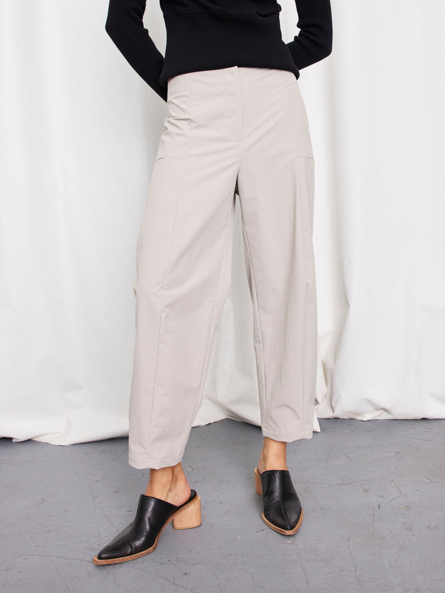 A person stands against a white curtain wearing the light beige Margot Pant by Zero + Maria Cornejo, paired with a black long-sleeve top and black slip-on mule shoes with a wooden heel. The person's face is not visible.