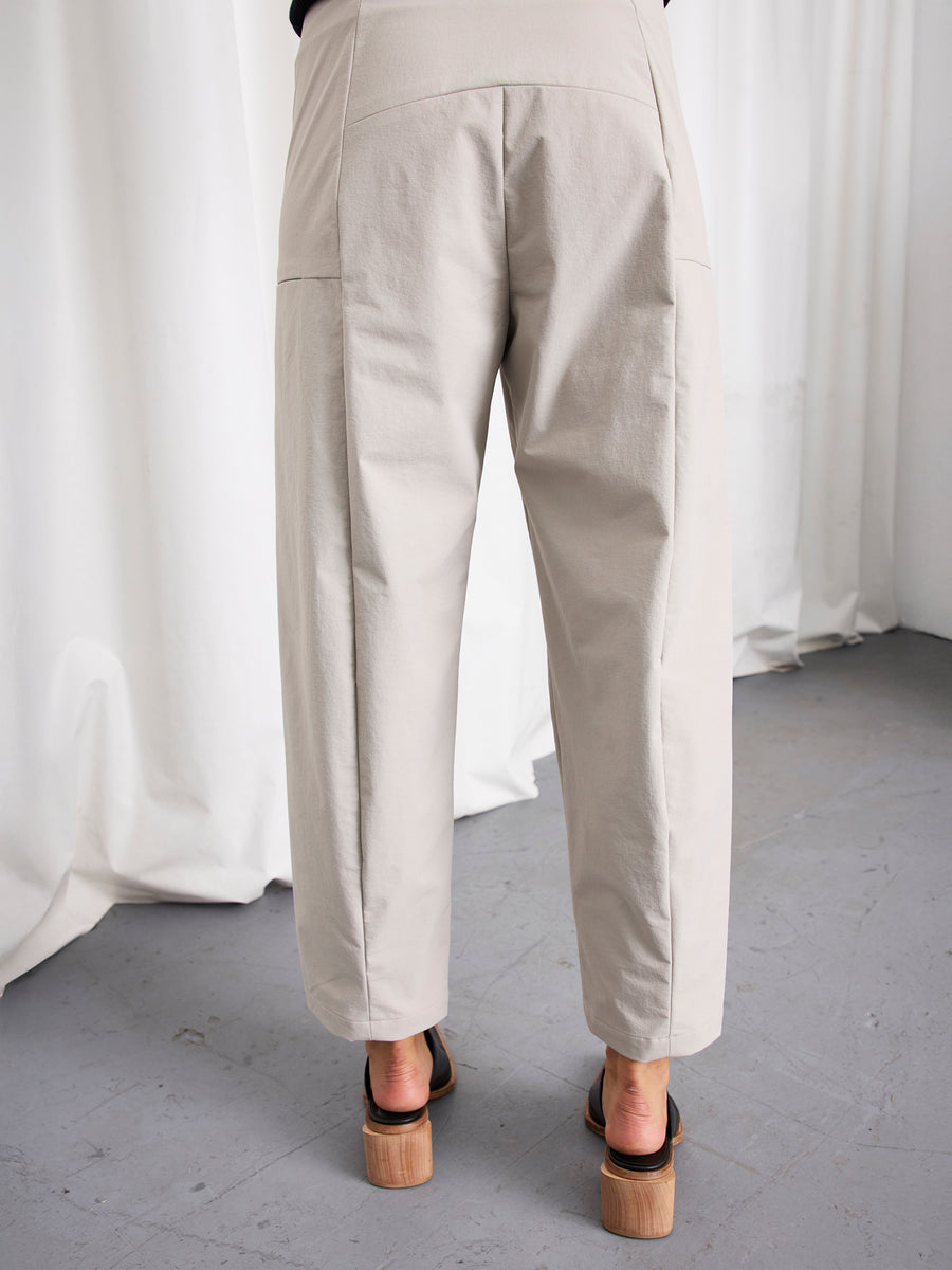A person stands on a gray floor wearing the Margot Pant by Zero + Maria Cornejo—loose-fitting, high-waisted, wide-leg cropped pants—paired with black chunky-heeled sandals. White curtains hang in the background.