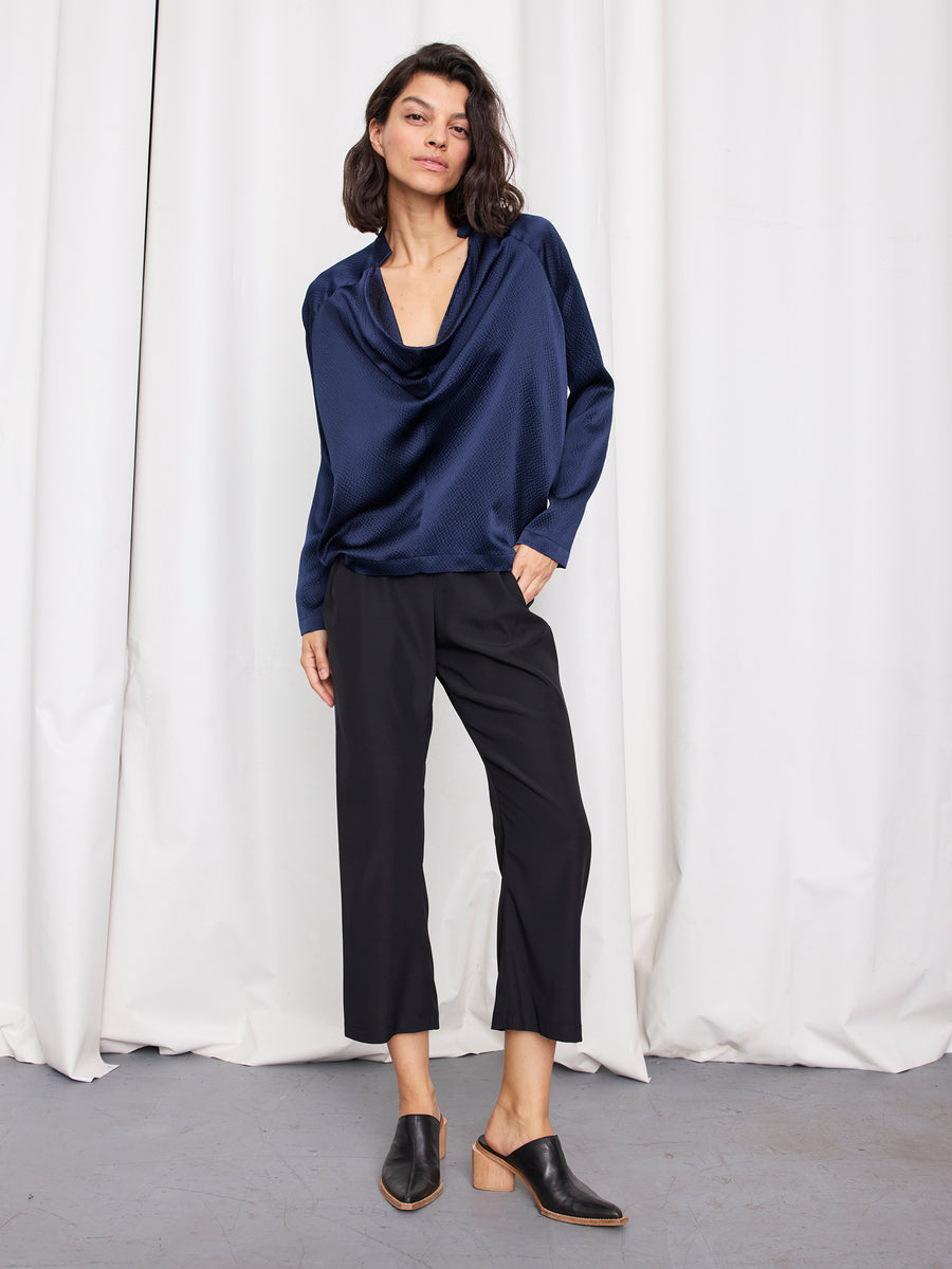A woman with dark shoulder-length hair poses confidently in front of white curtains, wearing the deep blue Long-Sleeved Evi Top by Zero + Maria Cornejo, paired with black cropped pants and black heeled mules.