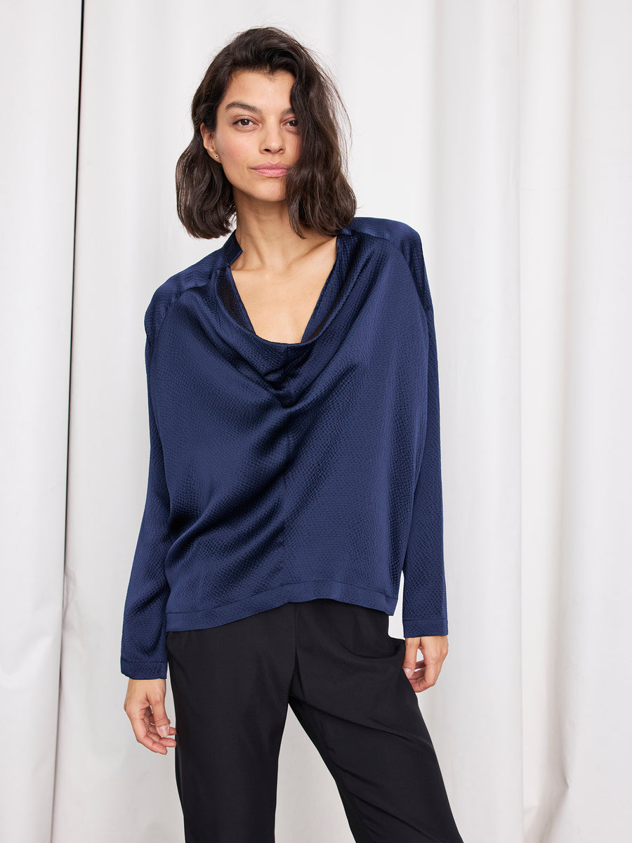 A woman with shoulder-length dark hair wears the Zero + Maria Cornejo Long-Sleeved Evi Top in deep blue sustainable silk and black pants, standing before a white curtain backdrop.