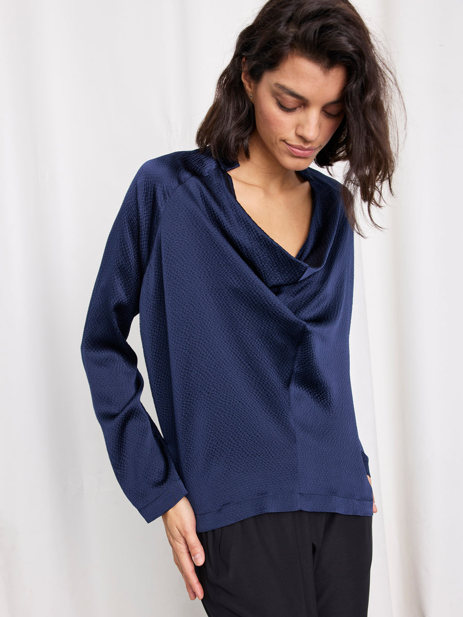A person with medium-length dark hair wears the Long-Sleeved Evi Top by Zero + Maria Cornejo, a loose-fitting, textured navy sustainable silk blouse with a draped neckline and long sleeves, paired with black pants against a white background.