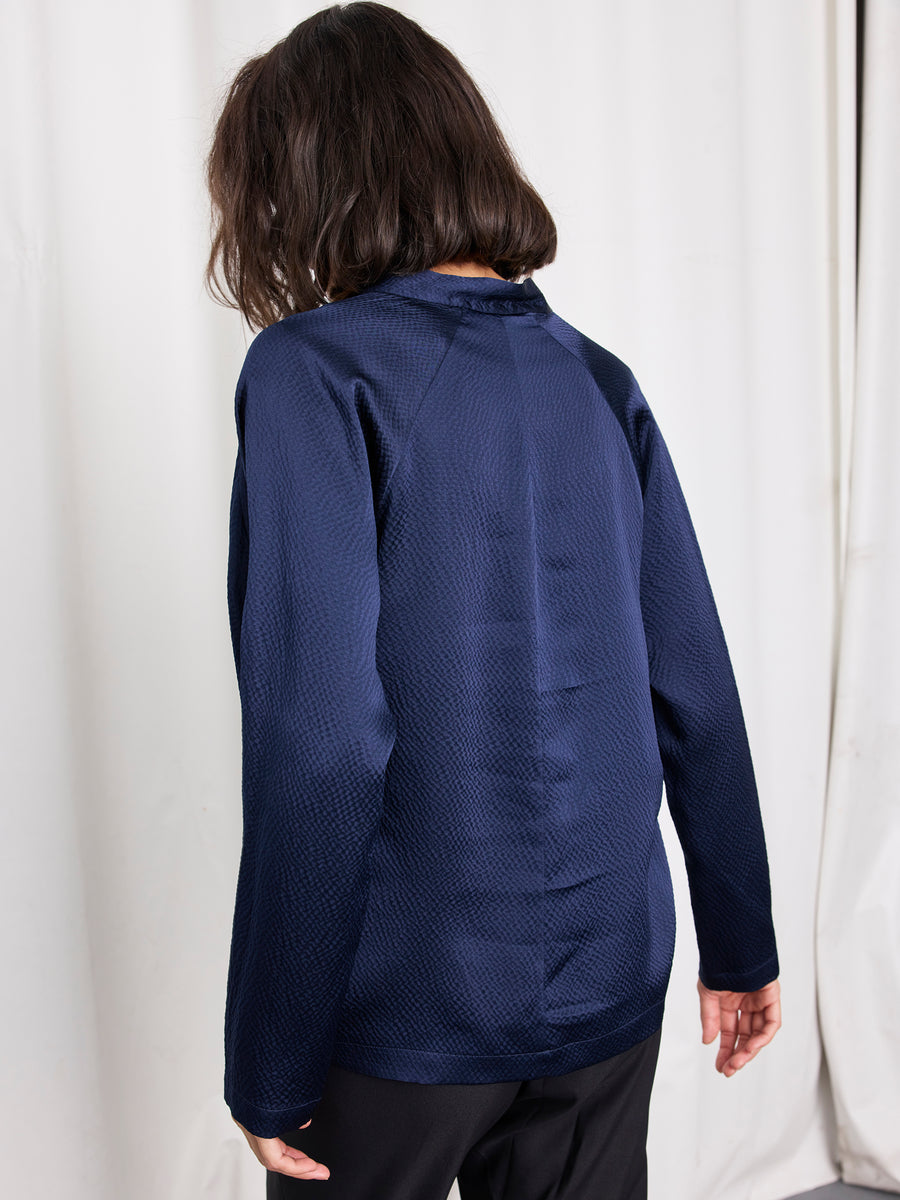 A person with shoulder-length brown hair, wearing the Zero + Maria Cornejo Long-Sleeved Evi Top in navy blue silk and black pants, stands facing away from the camera against a white curtain backdrop.