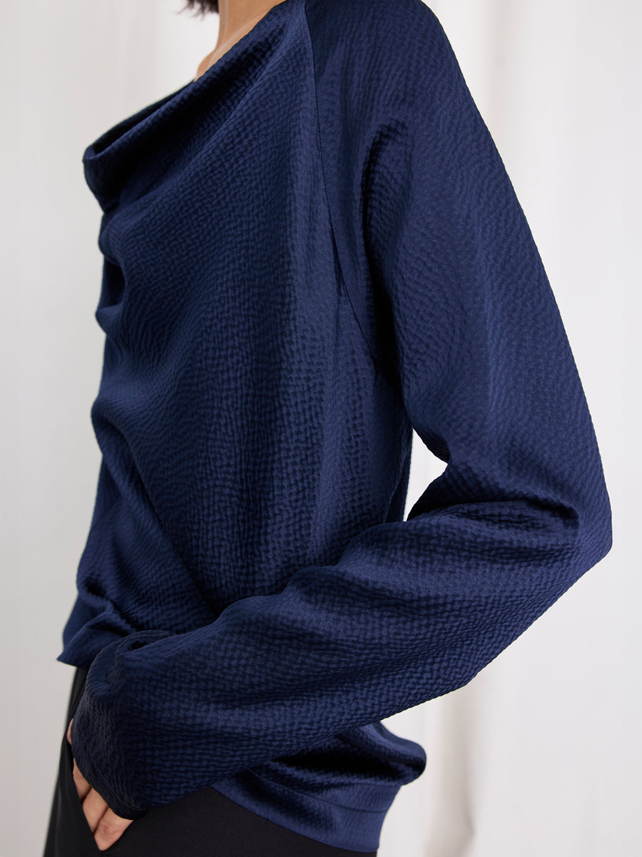 A person wears the Zero + Maria Cornejo Long-Sleeved Evi Top, a textured navy blue silk piece with a draped neckline. The shot is cropped to their torso and part of their face, set against a plain white background. Made in New York.