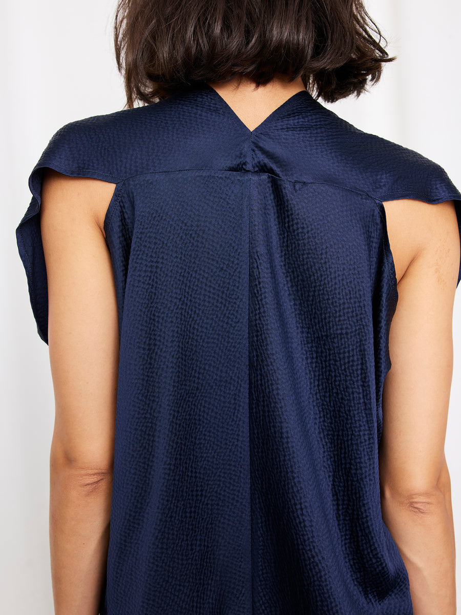 A person with short dark hair is shown from behind, wearing the Zero + Maria Cornejo Issa Dress—a textured dark blue, knee-length style featuring a distinctive V-shaped back neckline.
