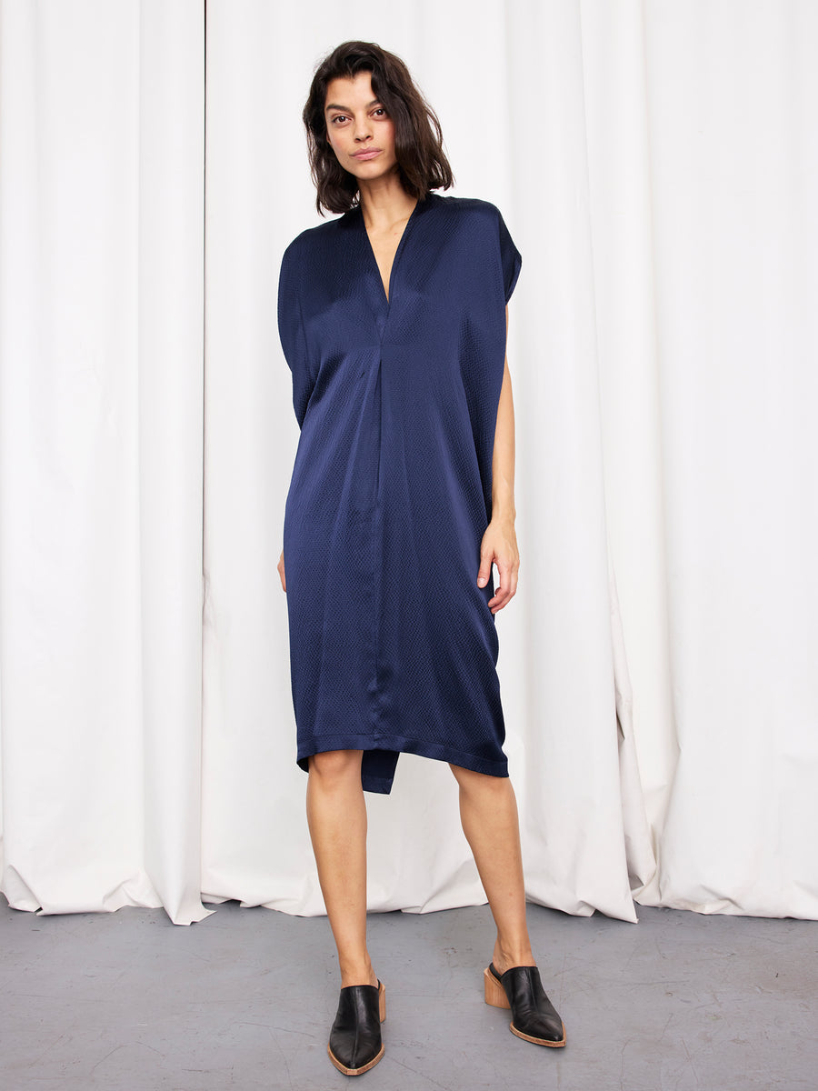 A woman stands before white curtains wearing the Issa Dress by Zero + Maria Cornejo—a loose-fitting, knee-length navy silk dress with short sleeves and a V-neck—paired with black mules for an effortlessly chic, sustainable look.