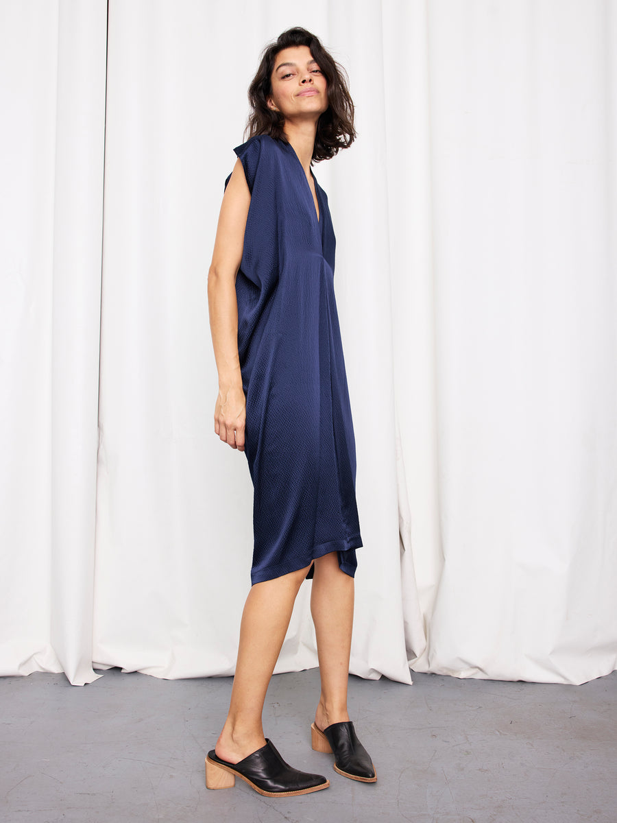 A woman poses in front of white curtains, wearing the Zero + Maria Cornejo Issa Dress, a loose-fitting, knee-length silk piece in deep blue. She completes her effortless sustainable look with black wooden-heeled mules.