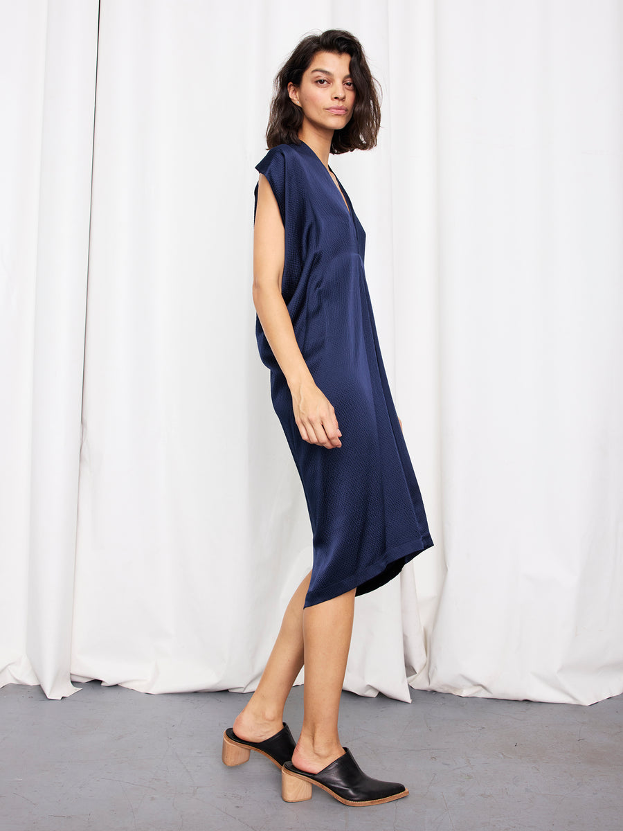 A person with shoulder-length dark hair wears the loose-fitting, sleeveless Issa Dress by Zero + Maria Cornejo and black low-heeled mules, standing sideways against a white curtain backdrop.