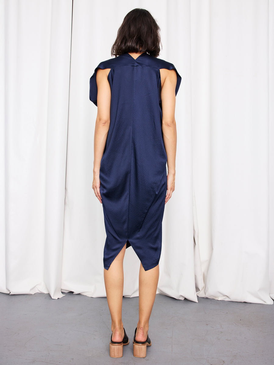 A woman faces away in the Issa Dress by Zero + Maria Cornejo—a loose, navy knee-length style with wide shoulders—styled with black heeled sandals, shoulder-length dark hair, and standing before white curtains for a sustainable fashion look.