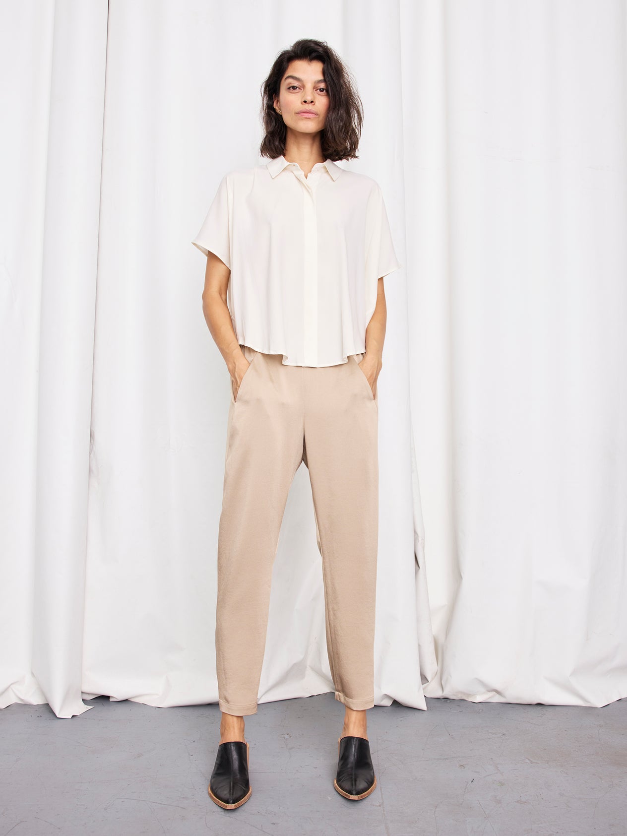 A person stands against a white wall wearing a black, loose-fitting, short-sleeved top made from recycled fabric and matching Gabi Trousers by Zero + Maria Cornejo with an elastic waist. They have long hair and are slightly leaning back with one hand in their pocket, complemented by black slip-on shoes.