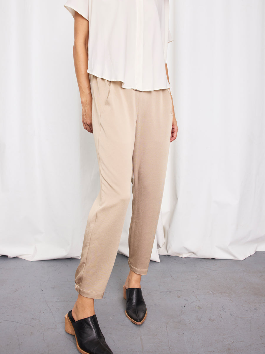A person stands against a white curtain on a gray floor, wearing Zero + Maria Cornejo Gabi Trousers in beige, made from recycled polyester, paired with a loose white short-sleeve top and black slip-on shoes.