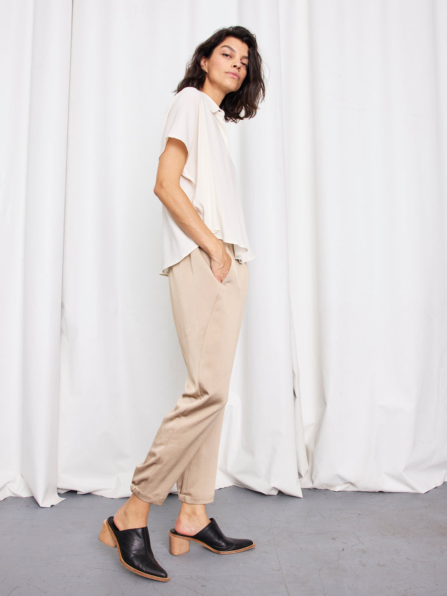 A person stands sideways against white curtains, wearing a loose white short-sleeve top, Zero + Maria Cornejo Gabi Trousers made in New York, and black heeled mules. One hand is in their pocket and they have a relaxed expression.