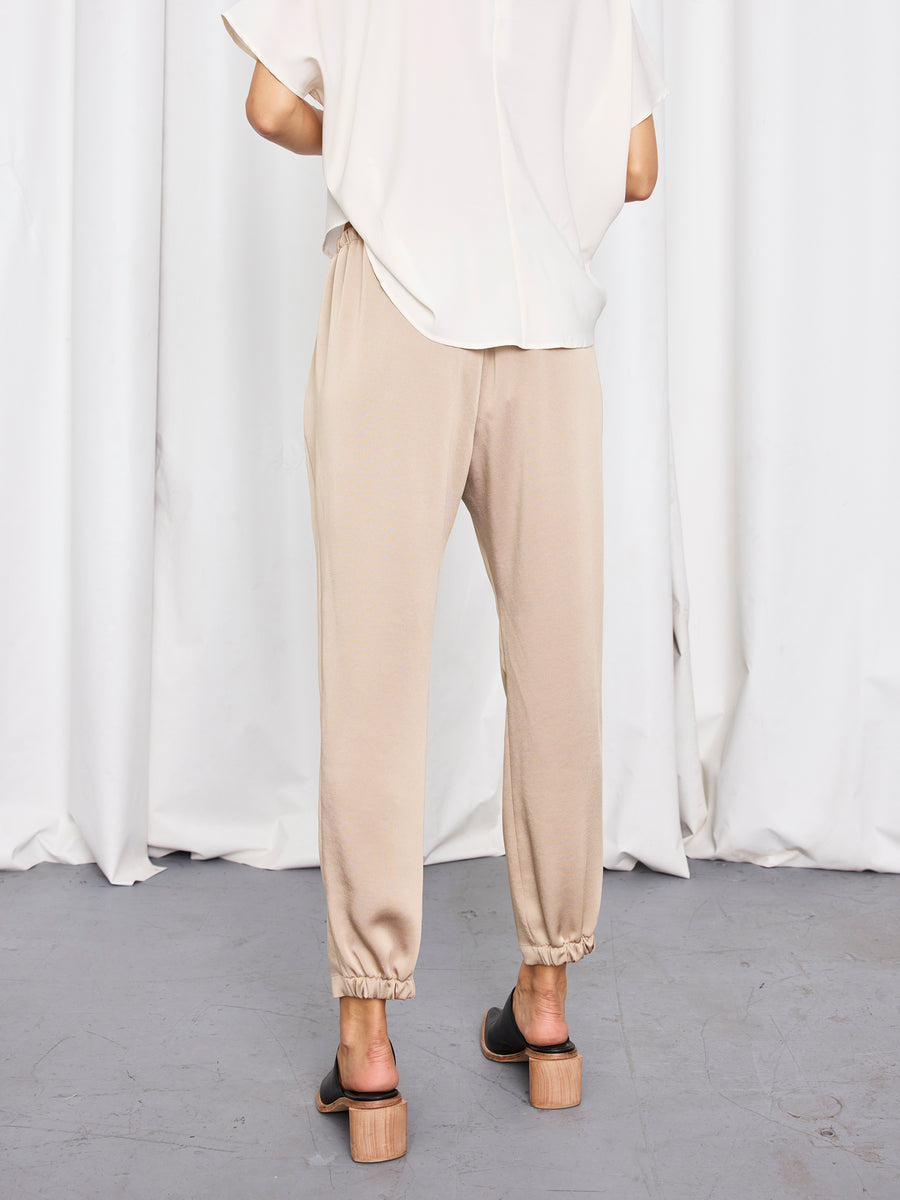 A person stands with their back to the camera, wearing a loose white short-sleeve top, Zero + Maria Cornejo’s Gabi Trouser made in New York, and black sandals with wooden block heels. A white curtain is in the background.