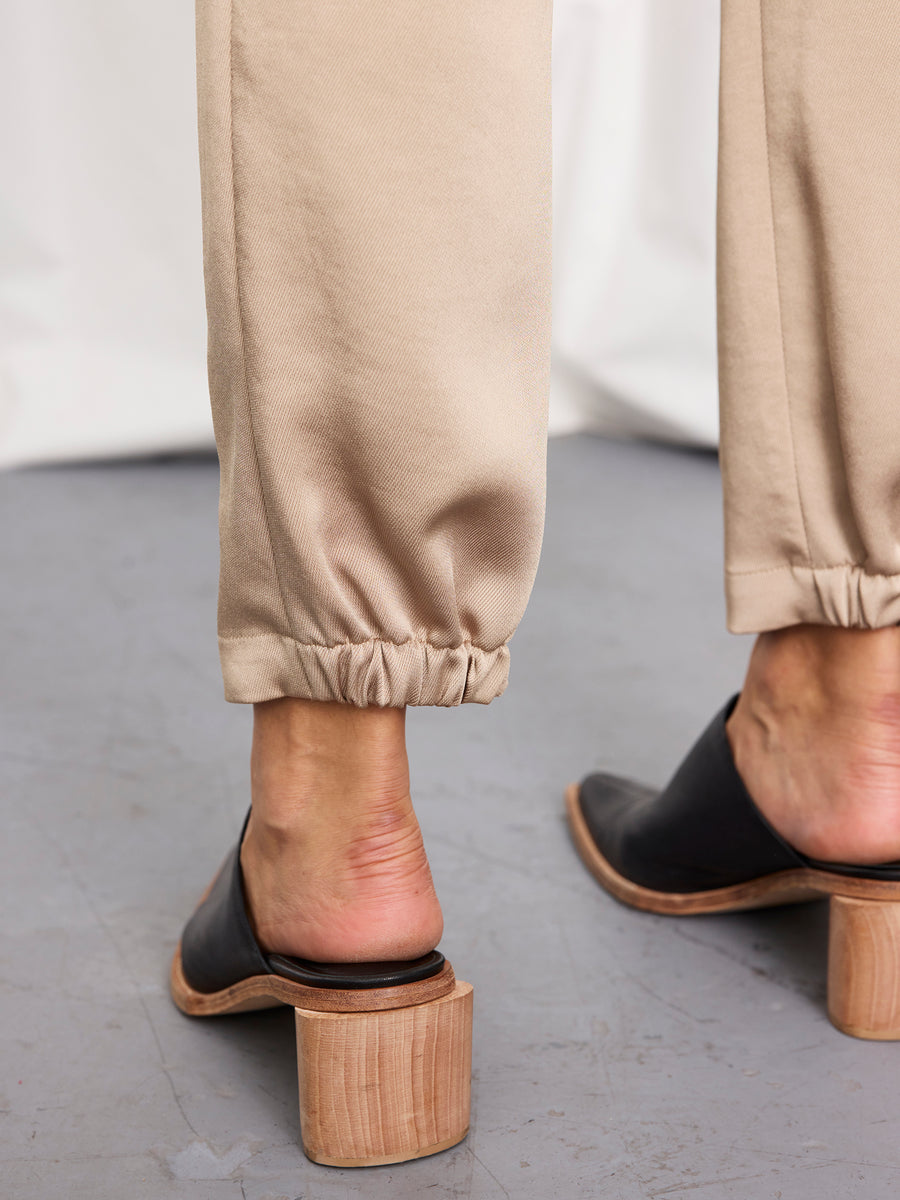 A close-up of a person’s lower legs in beige Zero + Maria Cornejo Gabi Trousers, featuring elastic cuffs and crafted from recycled drape fabric in New York, paired with black heeled mules on a gray floor.