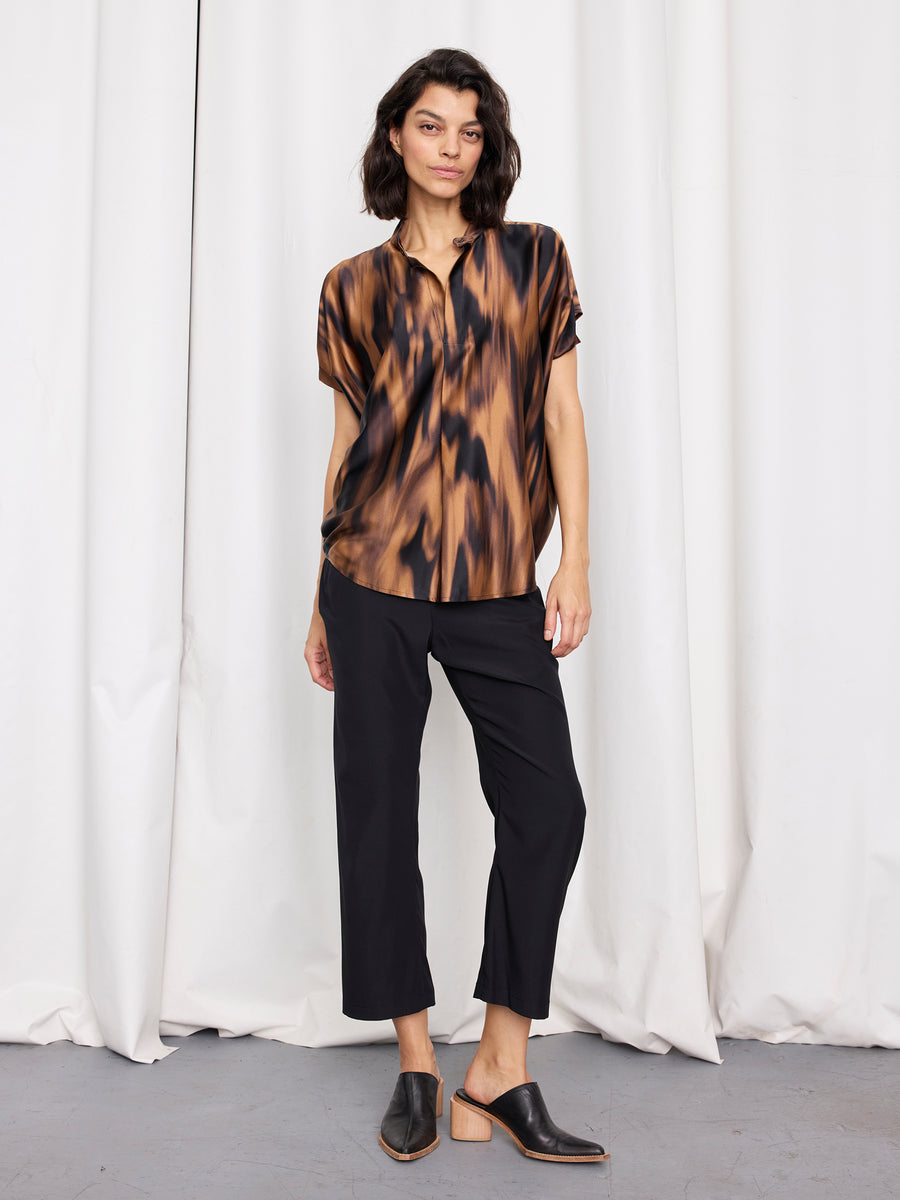 A woman stands before white curtains wearing the Mare Shirt by Zero + Maria Cornejo, featuring loose draped sleeves, paired with black cropped pants and black mule shoes with wooden heels.