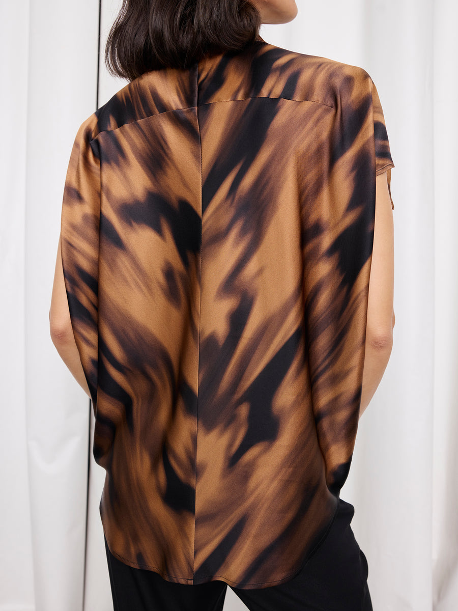 A person with shoulder-length dark hair is seen from behind wearing the Mare Shirt by Zero + Maria Cornejo, featuring a loose fit and brown and black abstract pattern, paired with black pants and standing before a light-colored curtain.