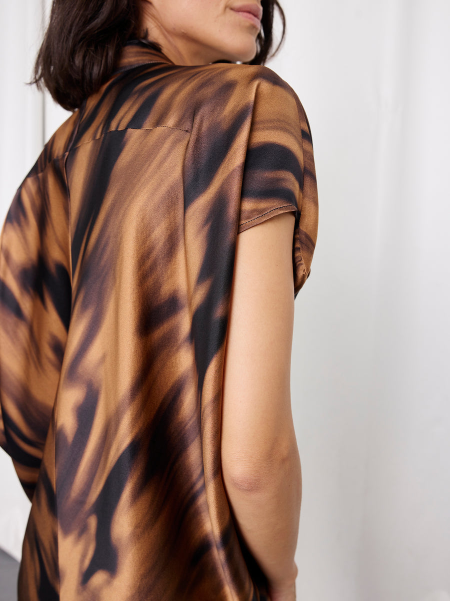 A person models the Zero + Maria Cornejo Mare Shirt—a loose, short-sleeve, brown and black patterned silk charmeuse top—standing sideways and backward against a plain white background with their face partially visible.