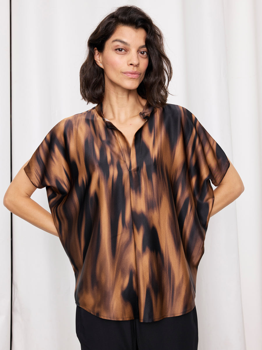 A woman with shoulder-length dark hair models the Mare Shirt by Zero + Maria Cornejo, a loose draped sleeve top with a brown and black abstract pattern, standing in front of white curtains, hands on her hips.