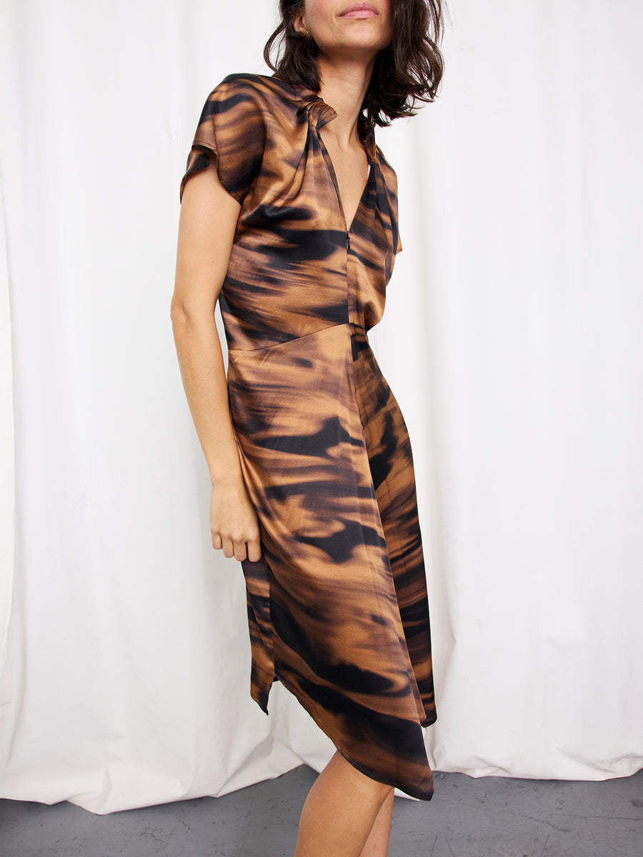 A woman wearing the Ruched Stella Dress by Zero + Maria Cornejo, a short-sleeved, knee-length silk charmeuse dress with a brown and black abstract pattern, stands before a white curtain with her dark hair loose and head slightly turned.