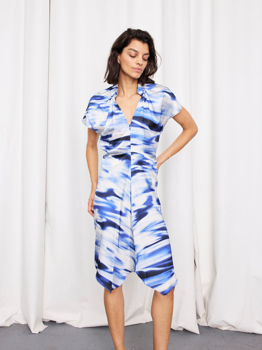A woman stands before white curtains wearing the Ruched Stella Dress by Zero + Maria Cornejo, featuring blue and white silk charmeuse with an abstract pattern, short sleeves, a draped hem, and one hand on her hip.