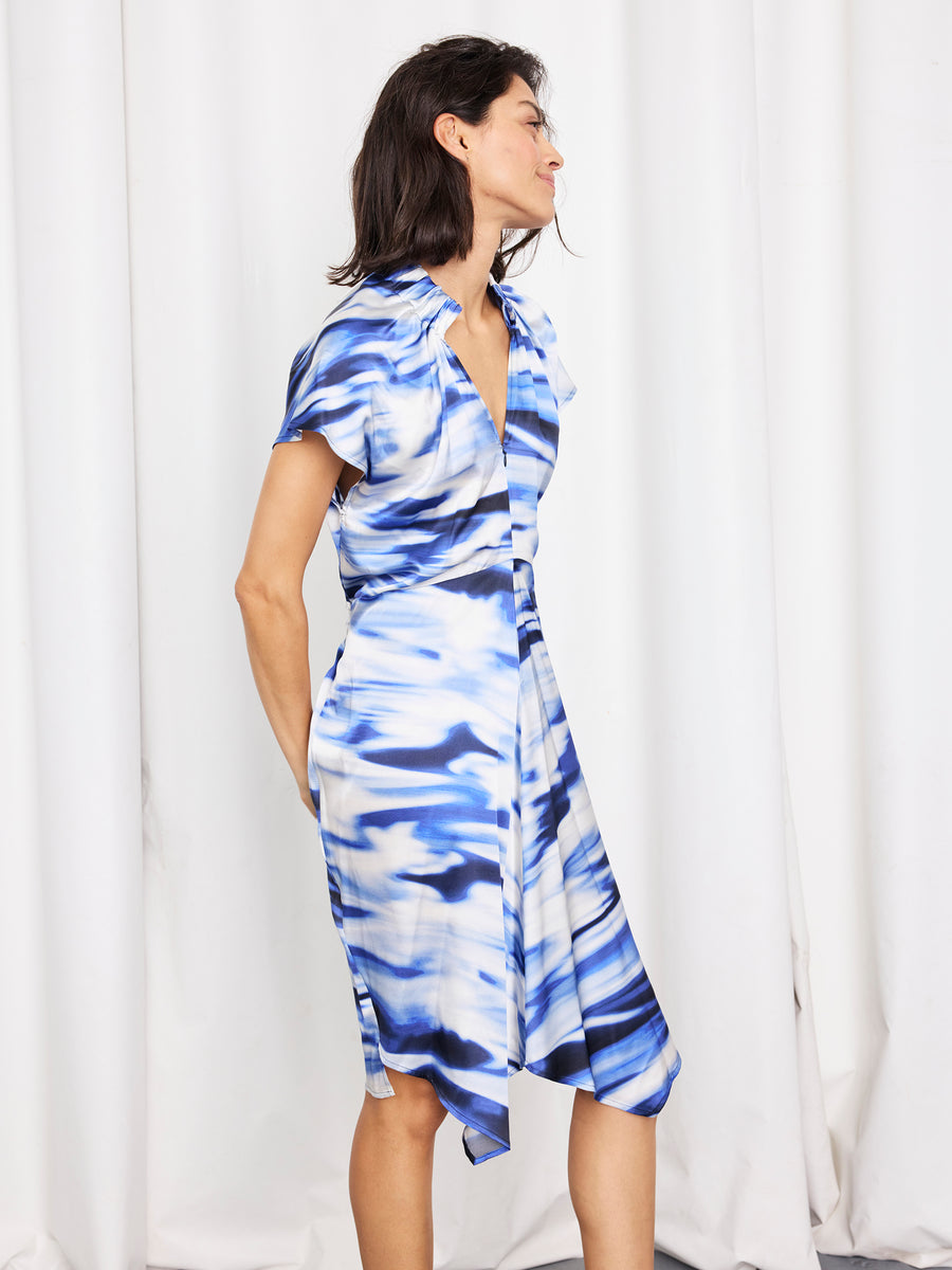 A woman stands sideways in front of white curtains, wearing the Ruched Stella Dress by Zero + Maria Cornejo—a knee-length, blue and white patterned silk charmeuse dress made in New York—with a hand in her pocket and a slight smile.