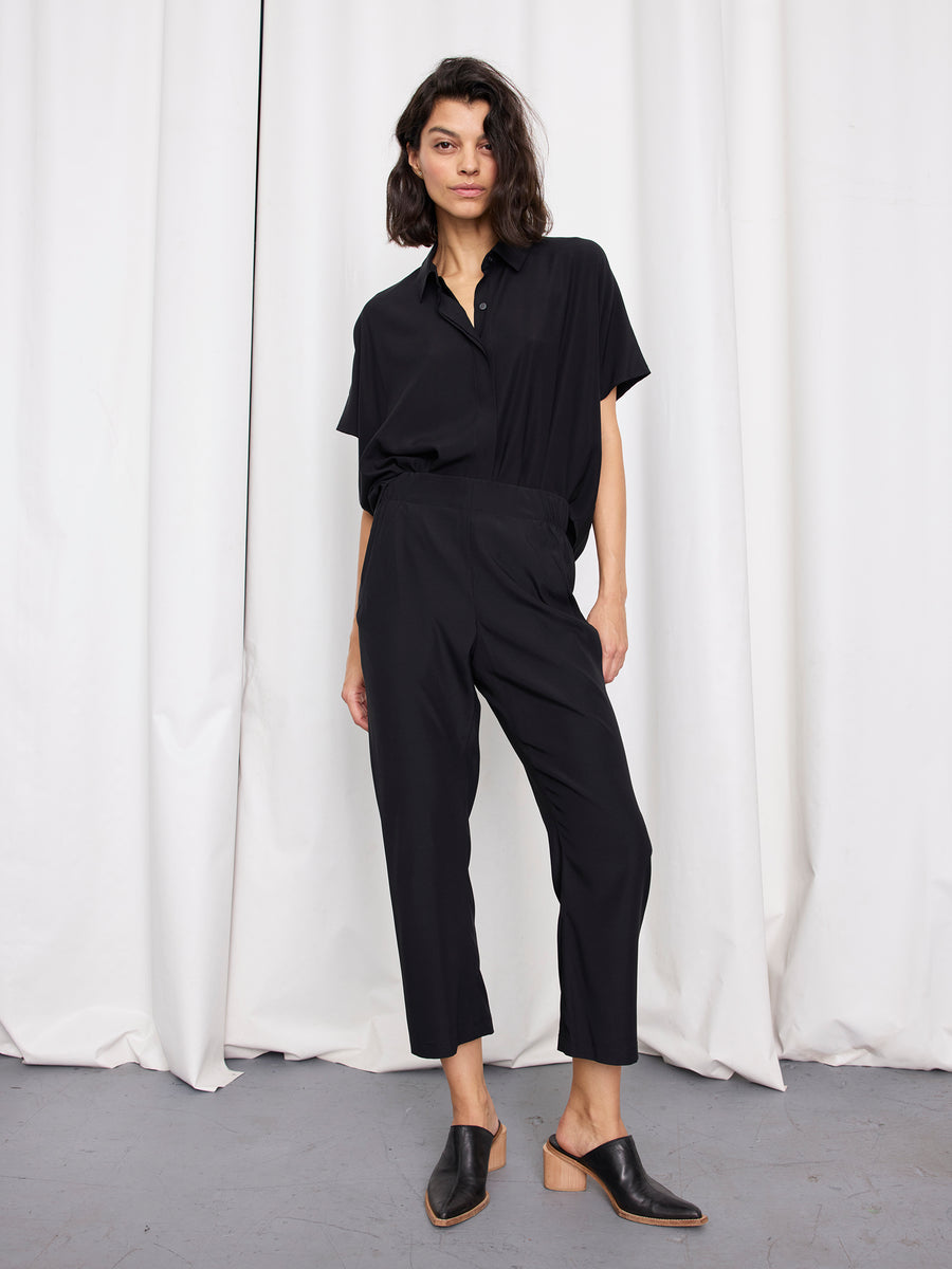 A person with shoulder-length dark hair wears a loose black short-sleeve shirt, Zero + Maria Cornejo Eko Pant in black, and black heeled mules, standing before white curtains on a gray floor.