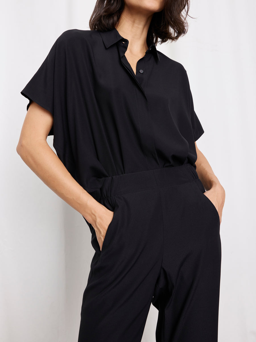 A person stands with hands in the pockets of Zero + Maria Cornejo's black, loose-fitting Eko Pant, paired with a short-sleeve, button-up black shirt. Their head is slightly turned against a plain white background.
