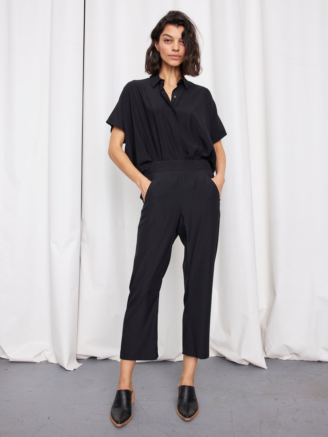 A woman stands confidently before white curtains, wearing Zero + Maria Cornejo’s loose-fitting black Eko Pant with her hands in her pockets—evoking effortless elegance.