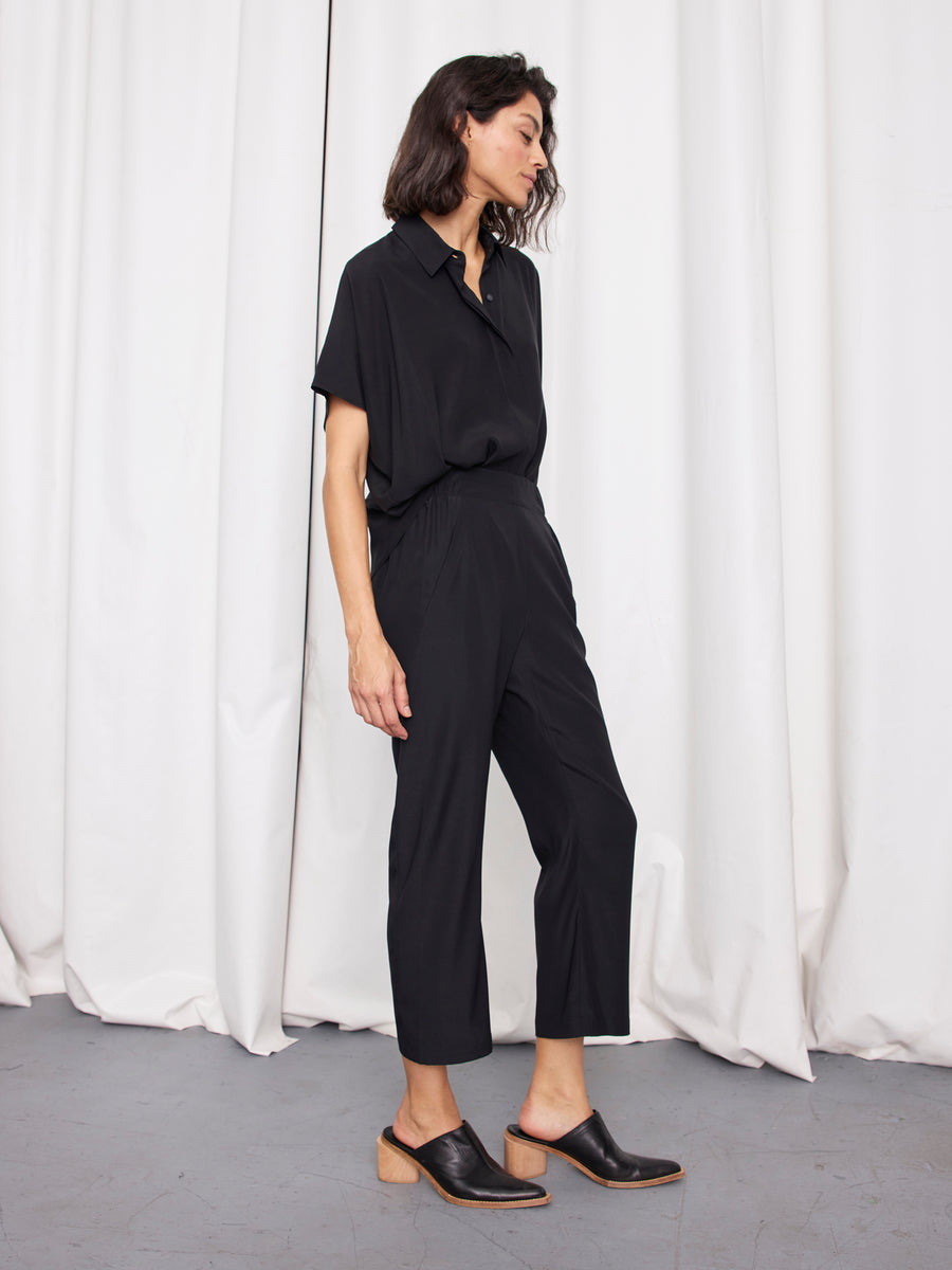 A woman with shoulder-length dark hair stands sideways in front of white curtains, wearing a black shirt tucked into the Eko Pant by Zero + Maria Cornejo and black mules with wooden soles.