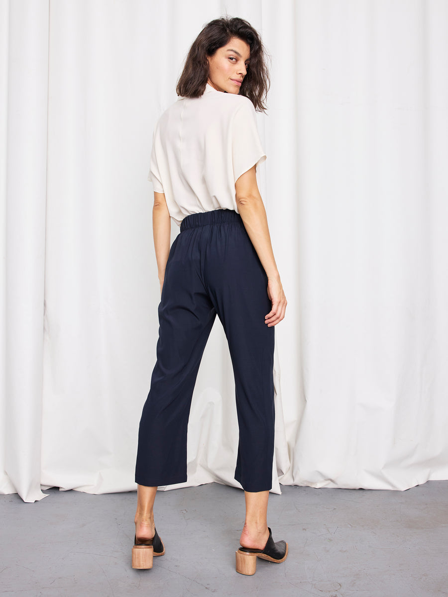 A woman stands indoors on a gray floor, facing backward. She wears a short-sleeved cream top, black heeled sandals, and the navy Eko Pant by Zero + Maria Cornejo. White curtains hang in the background.