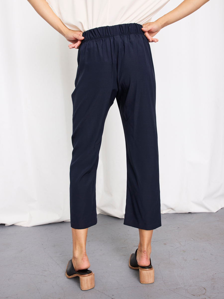 A person stands facing away, wearing the Zero + Maria Cornejo Eko Pant—navy charmeuse cropped trousers with an elastic waistband—paired with black heeled sandals, against a white curtain backdrop. Made in New York.