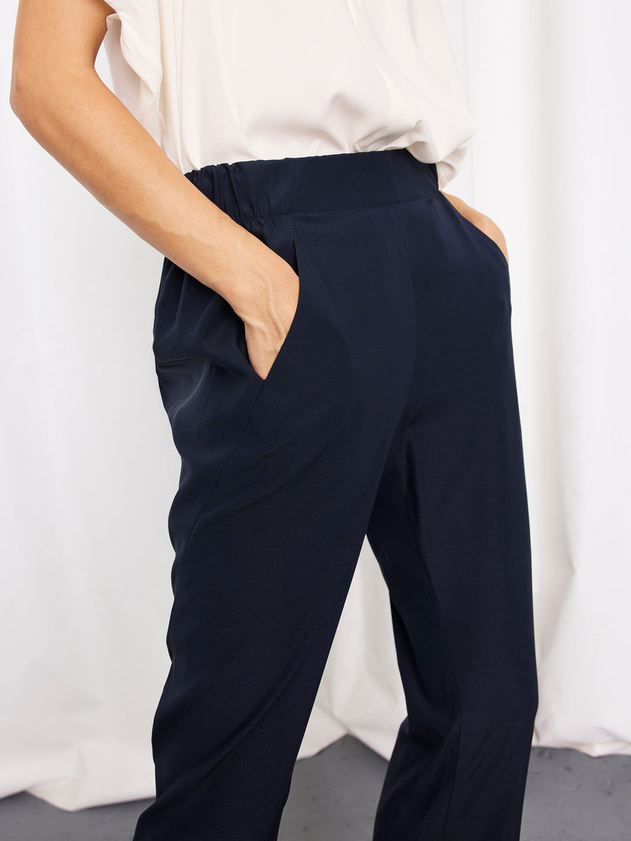A person stands with hands in the pockets of the high-waisted, dark navy Eko Pant by Zero + Maria Cornejo, paired with a loose white top. The background features soft draping fabric against a plain white setting.