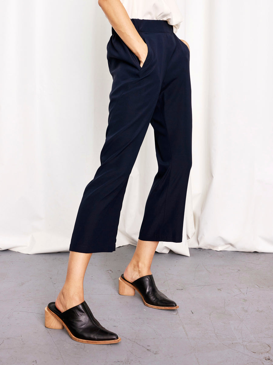 Person stands on a gray floor wearing Zero + Maria Cornejo Eko Pant in navy stretch silk, paired with a white top and black heeled mules with wooden soles, in front of a white curtain backdrop. Made in New York.