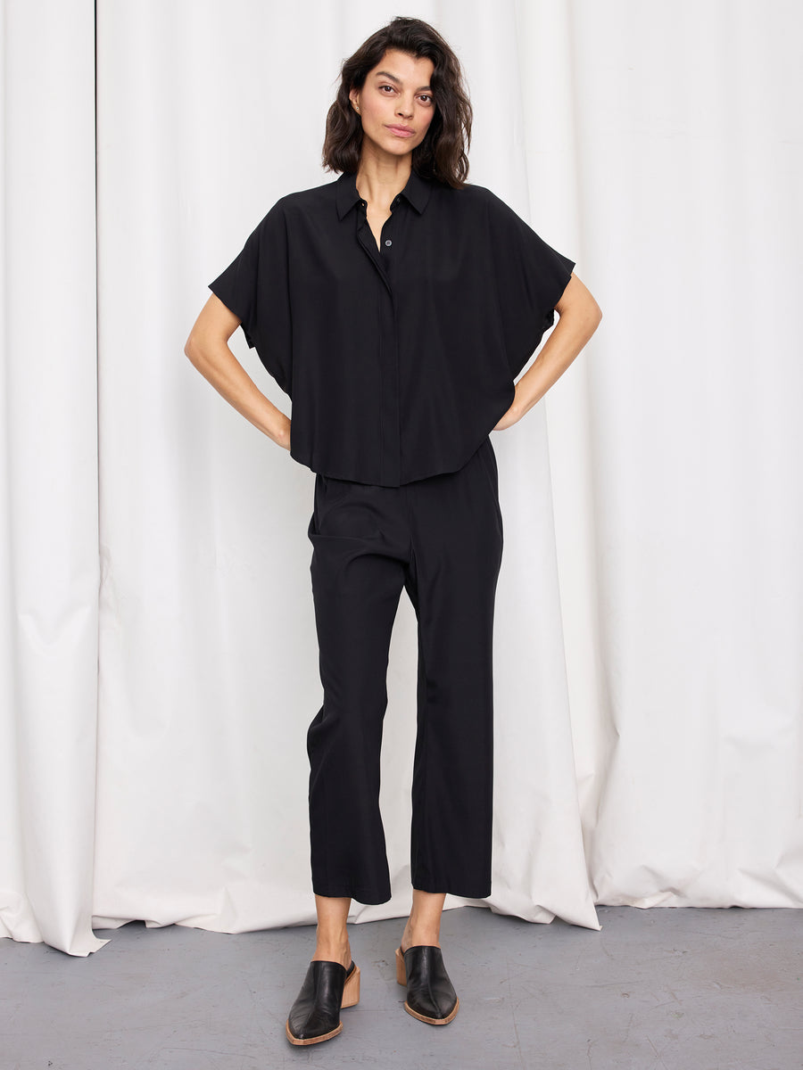 A person stands against white curtains, wearing the Circle Shirt by Zero + Maria Cornejo— a loose, short-sleeved silk top made in New York— with black cropped pants and slip-on shoes. One hand is in their pocket; their expression is relaxed.