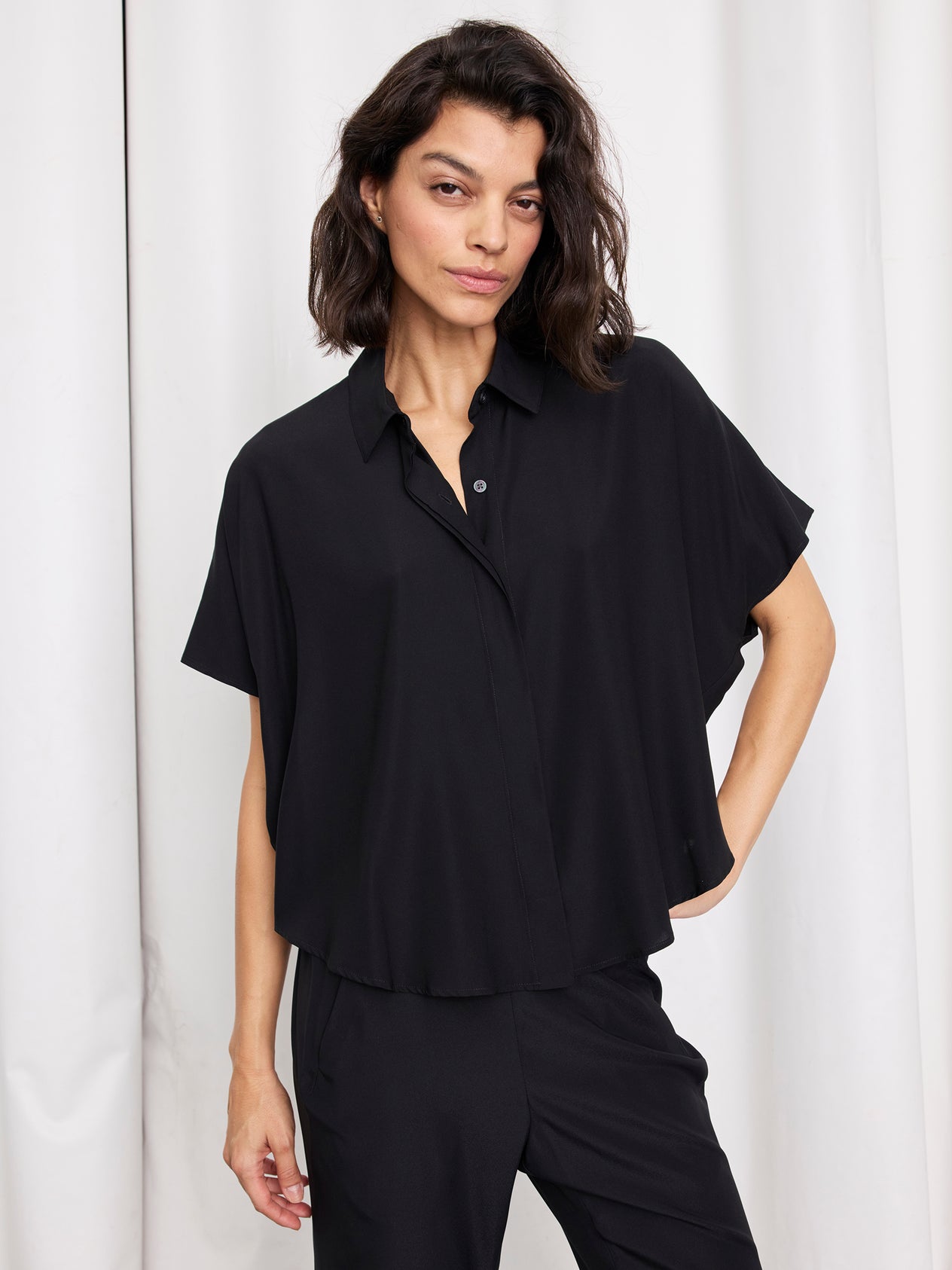 A woman with shoulder-length dark hair wears the loose-fitting Circle Shirt by Zero + Maria Cornejo with matching black pants, standing before white curtains with one hand on her hip.