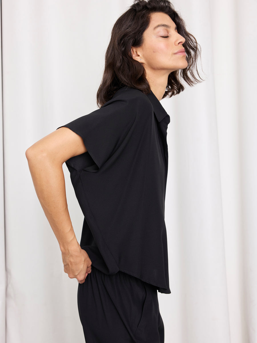 A person with medium-length dark hair stands in profile against a white curtain, eyes closed and hand on hip, wearing the Zero + Maria Cornejo Circle Shirt in black silk charmeuse and black pants.