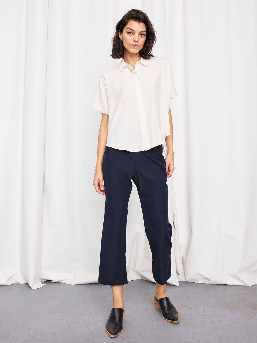 A person stands before white curtains, wearing the Zero + Maria Cornejo Circle Shirt, navy cropped pants, and black mules. With shoulder-length dark hair and a neutral expression, their look exudes effortless New York style.