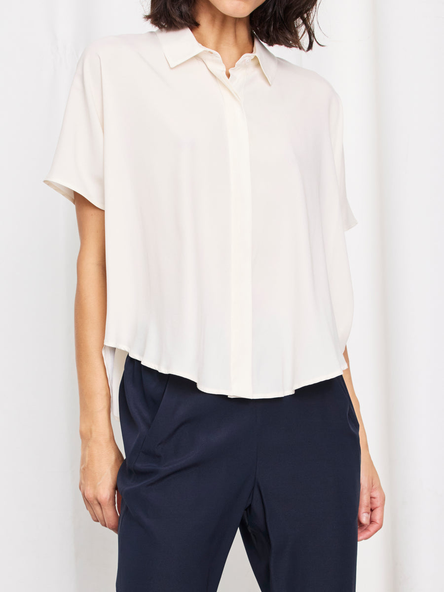 A woman wears the Zero + Maria Cornejo Circle Shirt—a loose silk charmeuse, short-sleeved collared top—paired with dark navy pants. Made in New York, the outfit is set against a white background, her face partially out of frame.