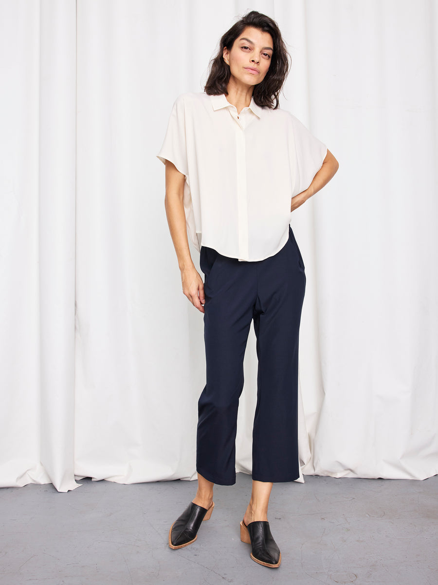 A person with shoulder-length dark hair stands indoors against white curtains, wearing a loose cream blouse, black mule shoes, and the Eko Pant by Zero + Maria Cornejo—navy stretch silk trousers made in New York. Their left hand rests on their hip.