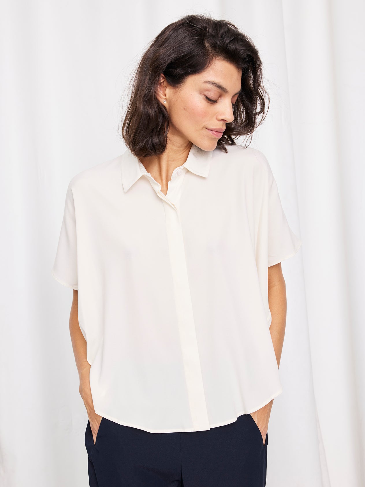 A woman with shoulder-length dark hair wears the loose-fitting Circle Shirt by Zero + Maria Cornejo with matching black pants, standing before white curtains with one hand on her hip.