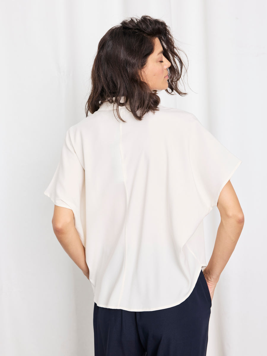 A woman with shoulder-length dark hair, hands in her pockets, models the loose-fitting Circle Shirt by Zero + Maria Cornejo with dark pants, standing against a white background and facing away from the camera.