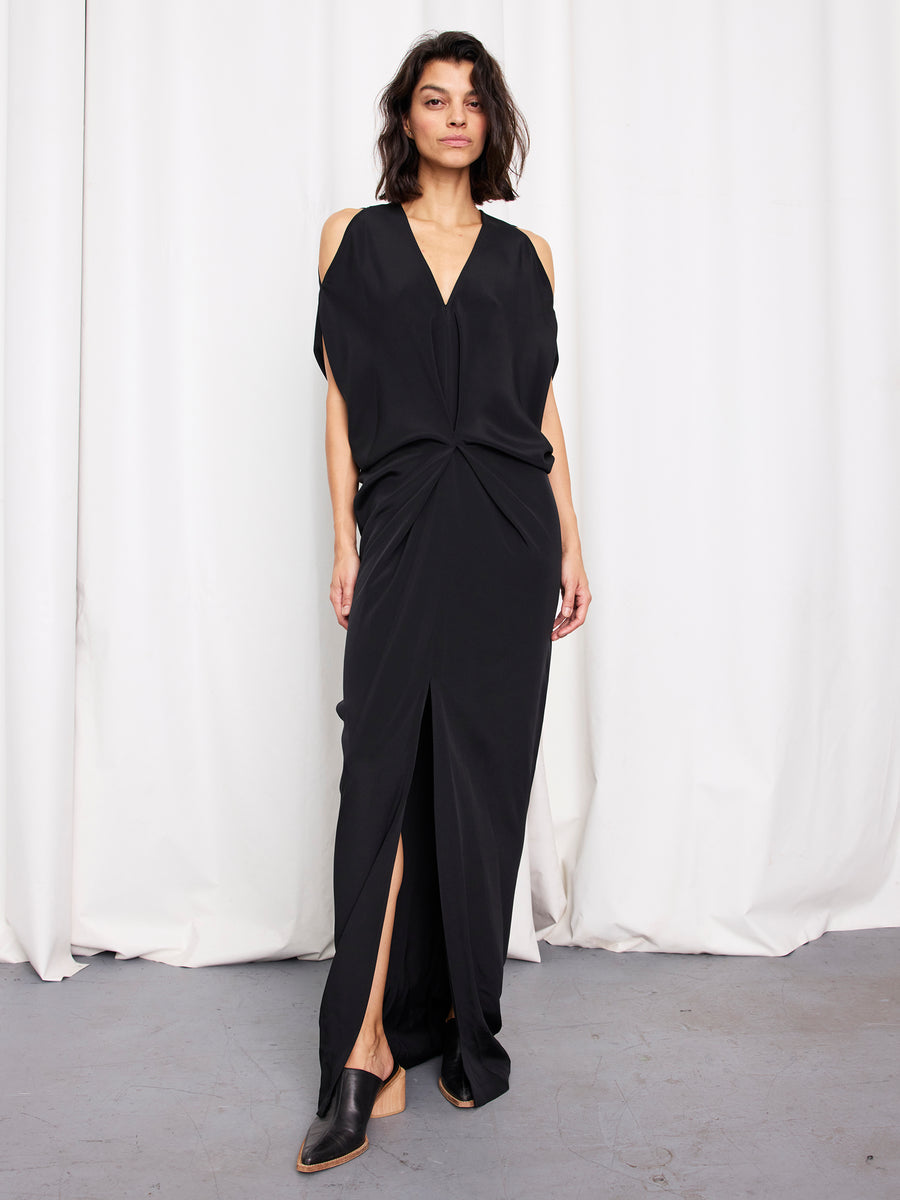 A woman stands before white curtains wearing the Long Miu Dress by Zero + Maria Cornejo—a black silk, full-length gown with cut-out shoulders, deep V-neckline, and high front slit—styled with black pointed-toe shoes. Made in New York.