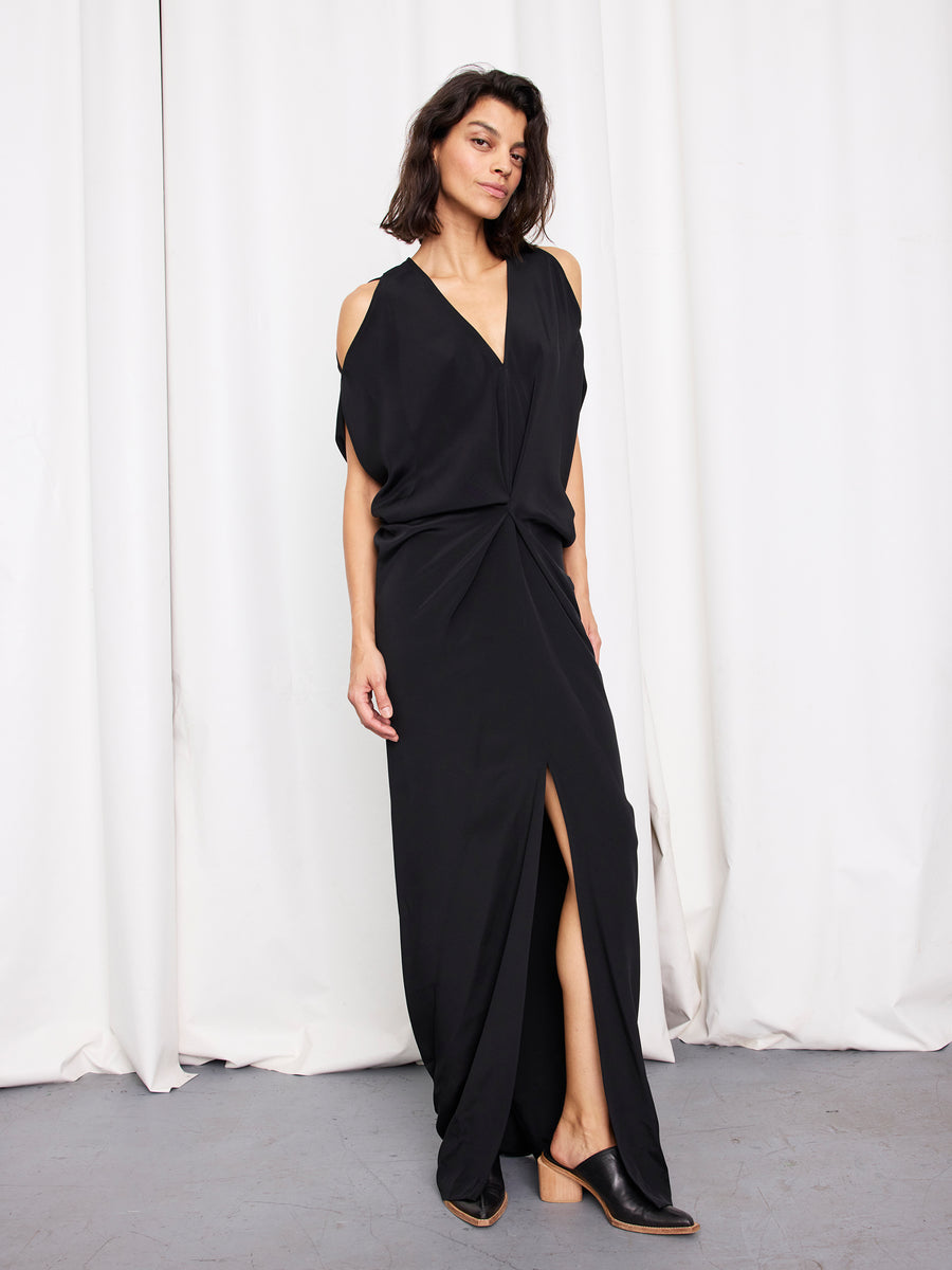 A woman wears the Long Miu Dress by Zero + Maria Cornejo—black, with cut-out shoulders and a front slit—standing on a gray floor before white curtains, paired with black slip-on shoes. Made in New York.